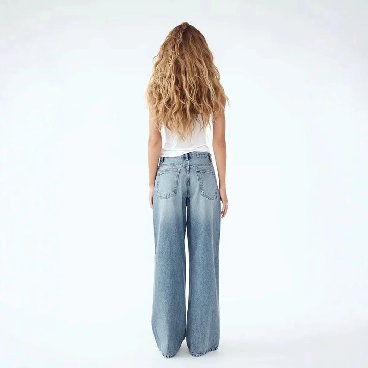 Woman with long wavy hair wearing white tank top and wide-leg blue jeans, standing back
