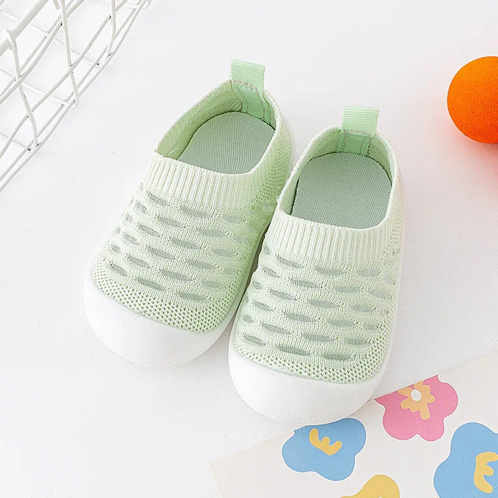Pair of light green knitted baby shoes with white soles on white surface with colorful paper and orange ball