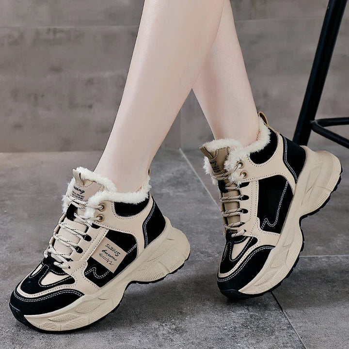 Beige and black chunky sneakers with fur lining worn on feet on a gray tiled floor