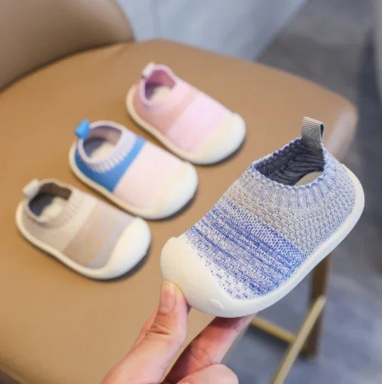 Hand holding a gray and blue knitted baby shoe with two pairs of pink and blue knitted baby shoes on chair