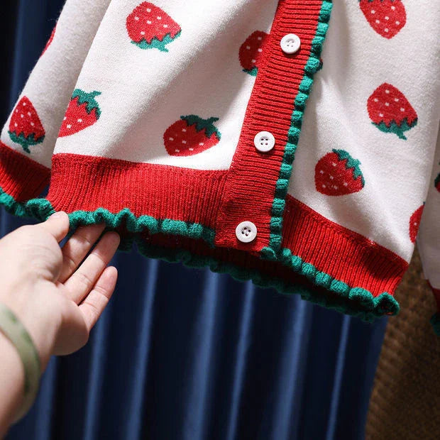 Close-up of a white cardigan with red strawberries, red trim, green ruffled edges, and white buttons on a blue background