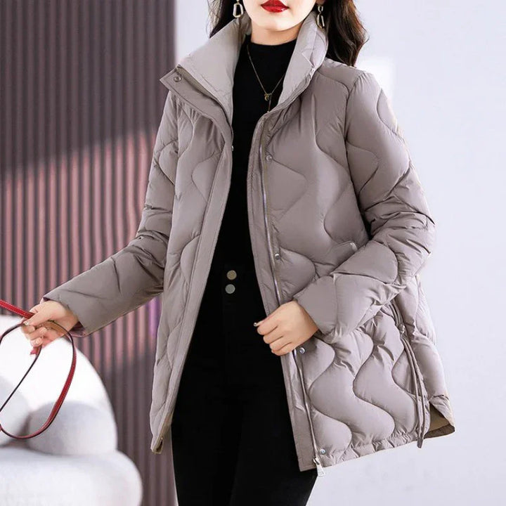 Woman wearing light gray quilted puffer jacket with high collar and zipper, black outfit underneath