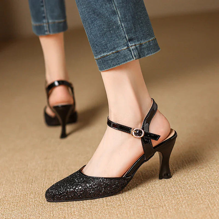 Black glitter pointed toe heels with ankle strap and buckle worn with cuffed jeans on beige carpet