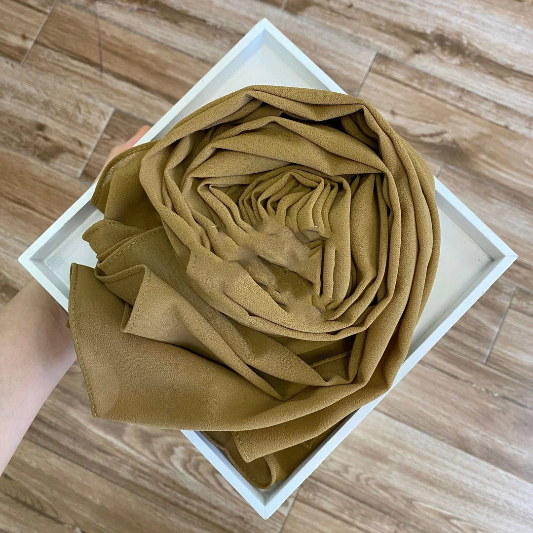 Person holding a white tray with a neatly rolled olive green chiffon scarf on wooden floor background