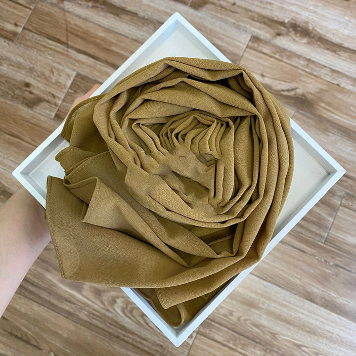 Person holding a white tray with a neatly rolled olive green chiffon scarf on wooden floor background