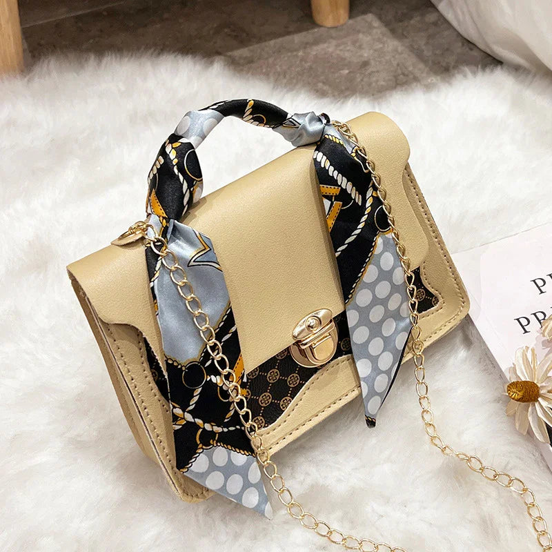 Beige leather handbag with gold clasp, chain strap, and black patterned silk scarf on white furry surface