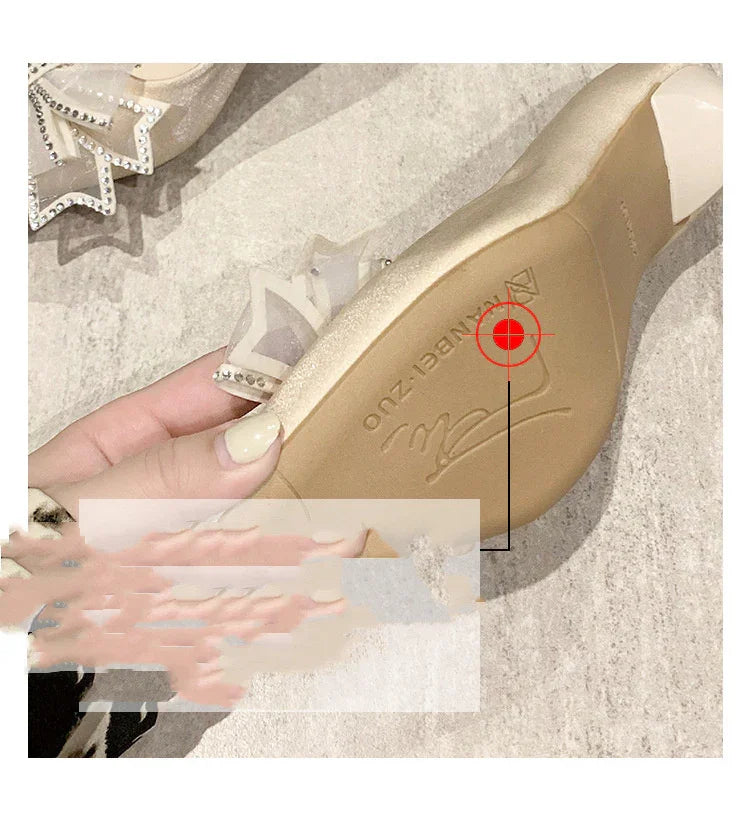 Hand holding beige flat shoe with transparent crystal-embellished bow detail on stone floor