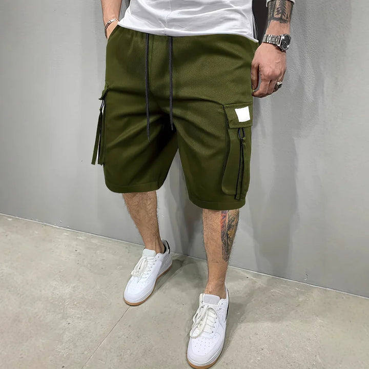 Man wearing olive green cargo shorts paired with white sneakers in a neutral indoor setting