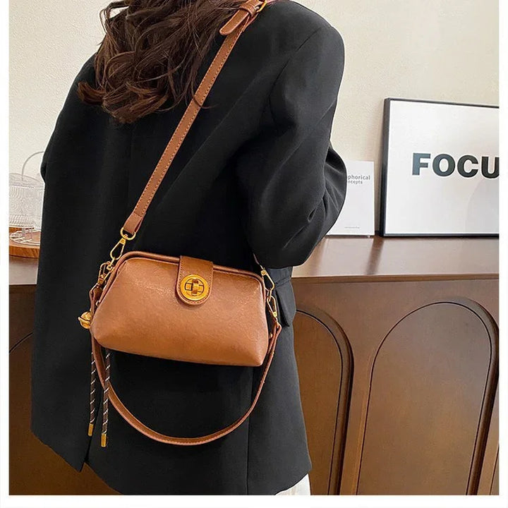 Brown leather crossbody bag with gold clasp worn over black blazer indoors