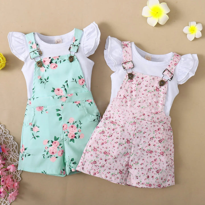 Two toddler floral overalls with white ruffled sleeve shirts, one mint green and one pink, on beige background