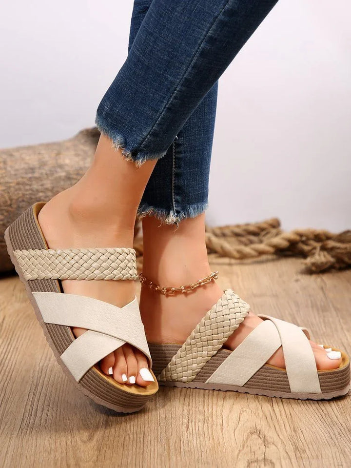 Close-up of feet in woven cream sandals with crisscross straps and gold anklet on wooden floor