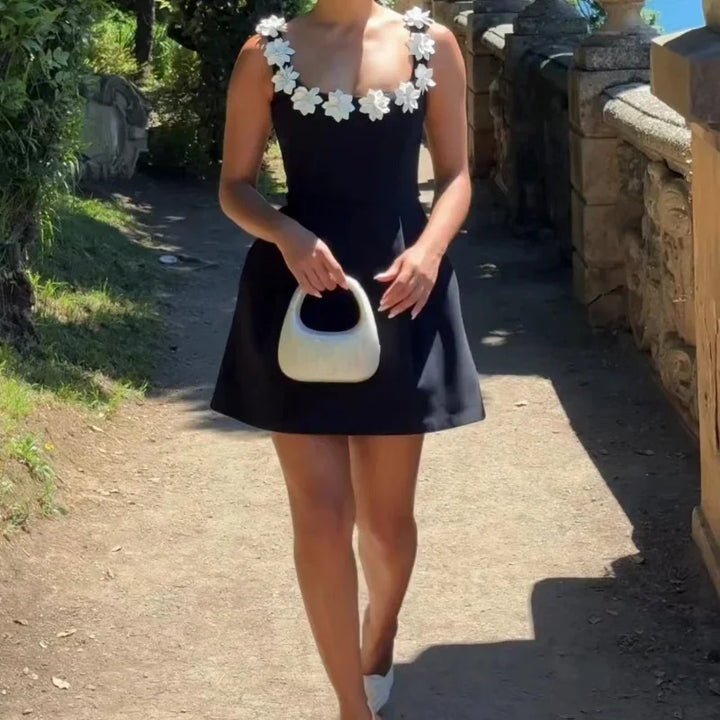 Woman wearing black sleeveless dress with white floral neckline holding white handbag outdoors