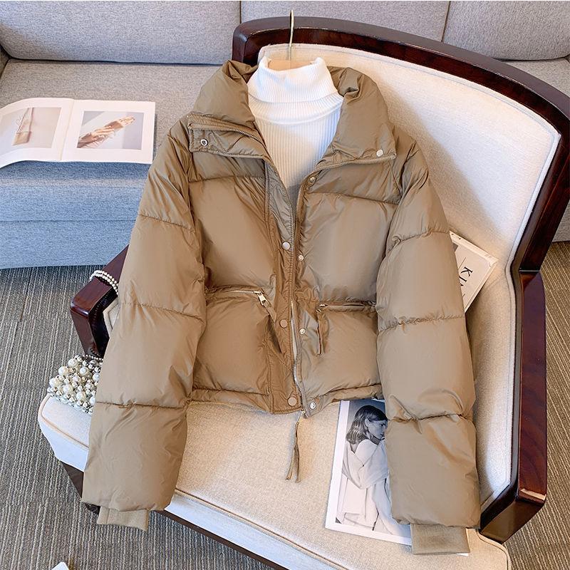 Beige puffer jacket with zipper and snap buttons displayed on a beige cushioned chair over white turtleneck sweater
