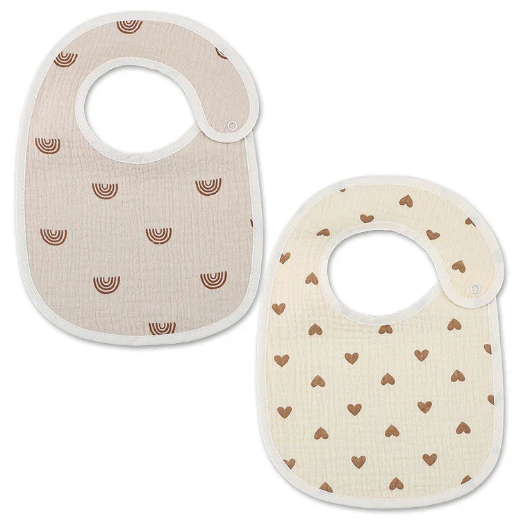 Two soft baby bibs with snap closures, one beige with brown rainbows, the other cream with brown hearts