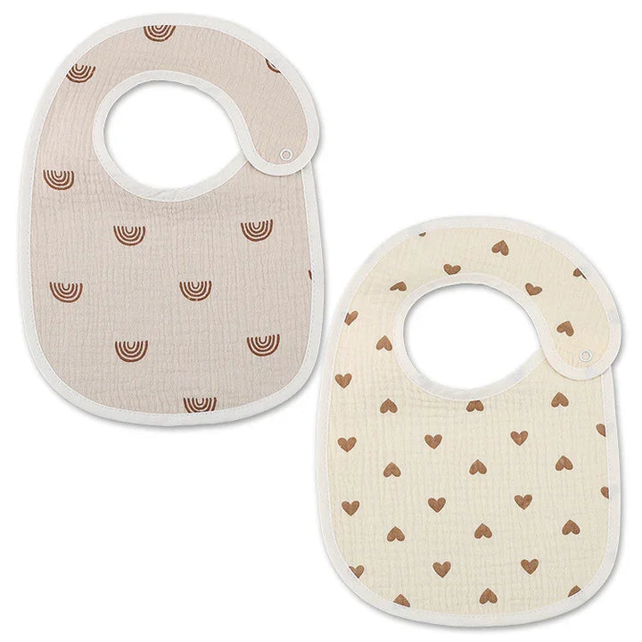 Two soft baby bibs with snap closures, one beige with brown rainbows, the other cream with brown hearts