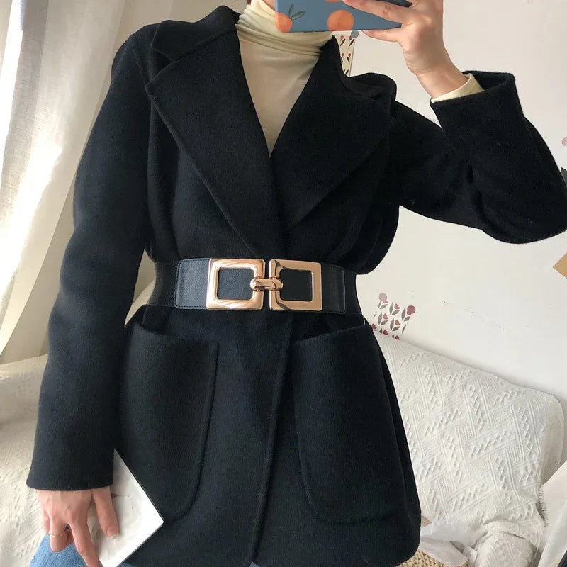 Woman wearing black wool coat with wide lapels and large gold buckle belt, cream turtleneck underneath