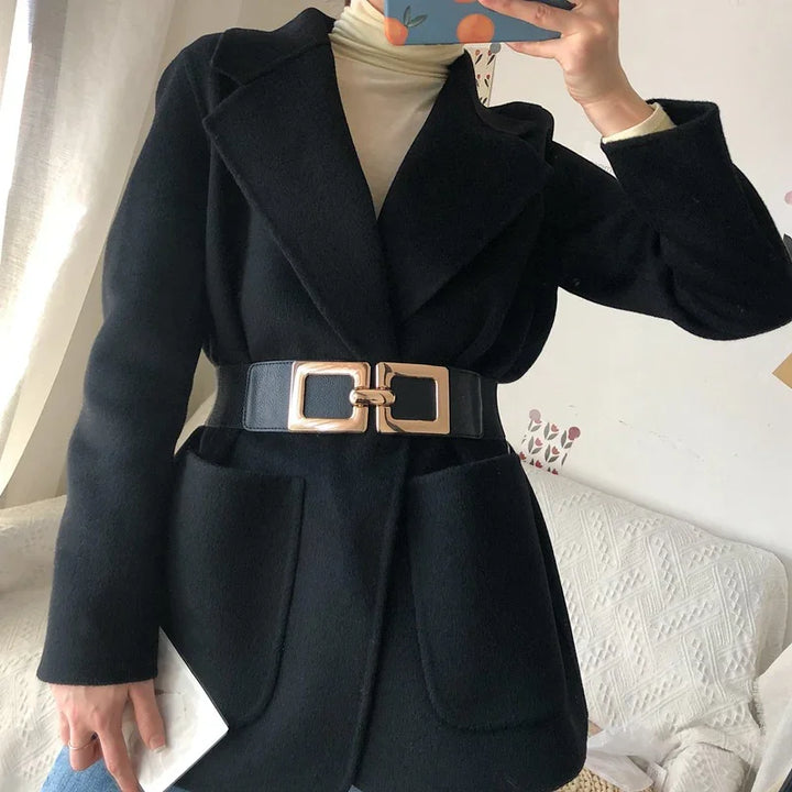 Woman wearing black wool coat with wide lapels and large gold buckle belt, cream turtleneck underneath