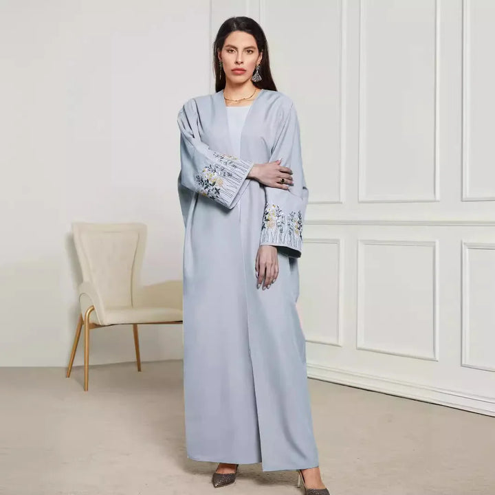 Woman in light blue abaya with floral embroidery on sleeves standing in minimalist room with cream chair