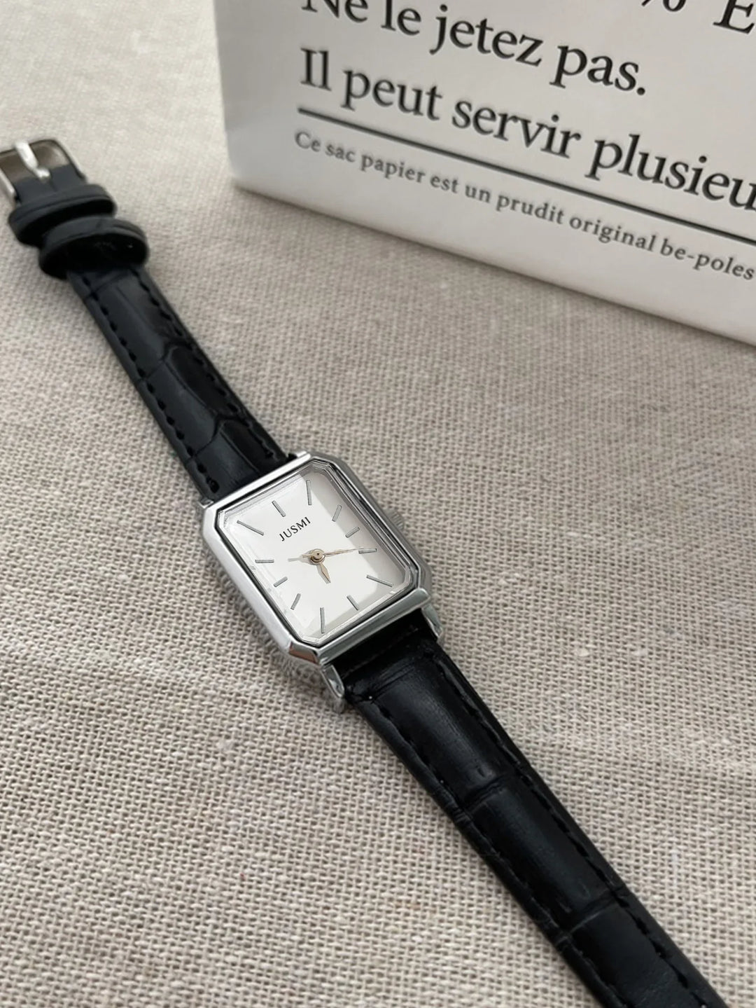 Rectangular silver wristwatch with black leather strap on beige fabric background near white paper bag