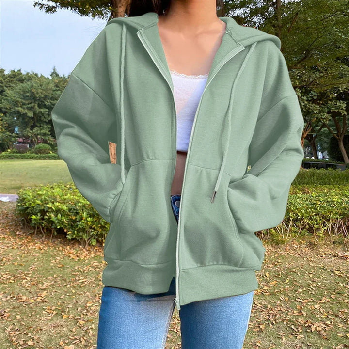 Woman wearing light green zip-up hoodie with front pockets and drawstrings, paired with white tank top and blue jeans outdoors in park