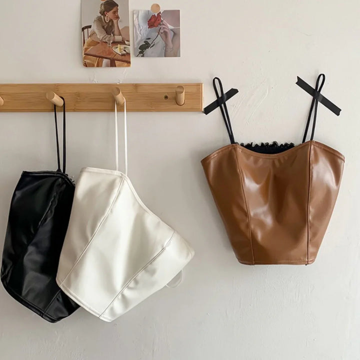 Three leather corset tops in black, white, and brown hanging on wooden hooks against a white wall