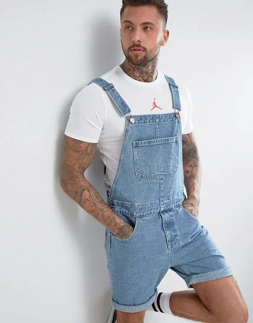man with tattoos wearing denim short overalls and white Jordan t-shirt leaning against white wall