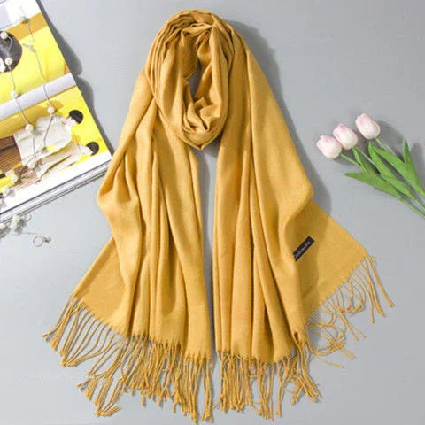 Soft mustard yellow scarf with fringes displayed on a gray surface next to tulips and a magazine