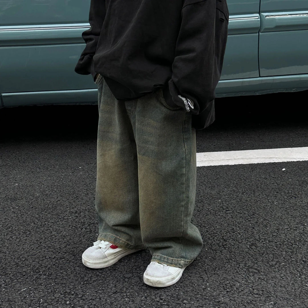 Person wearing oversized faded green jeans and white sneakers standing on asphalt near a light blue car