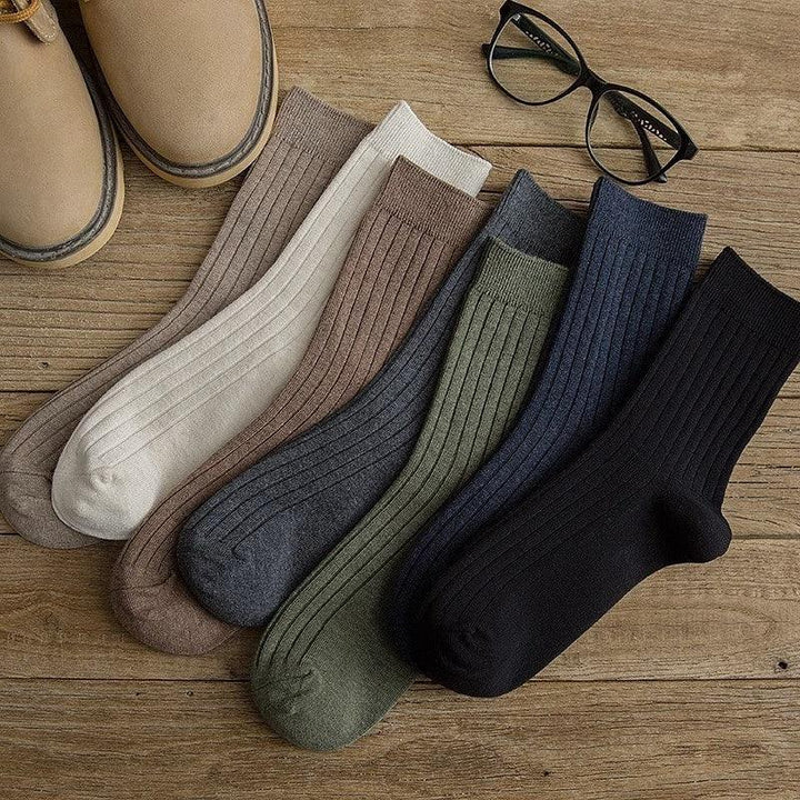 Set of seven ribbed cotton socks in neutral colors on wooden surface with brown boots and black glasses