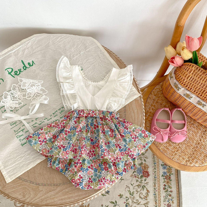 Floral baby dress with white ruffled top on wooden tray, pink baby shoes on chair with woven basket and tulips