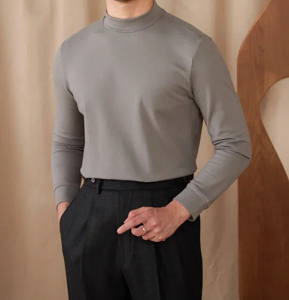 Man wearing a grey mock neck long-sleeve shirt tucked into high-waisted black pants standing indoors