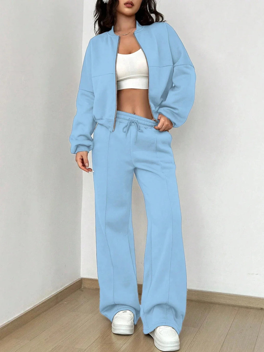 Woman wearing light blue zip-up jacket and matching wide-leg pants with white crop top and white sneakers indoors