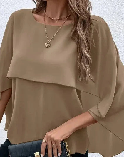 Woman wearing beige asymmetrical layered top with flowy sleeves and gold heart pendant necklace