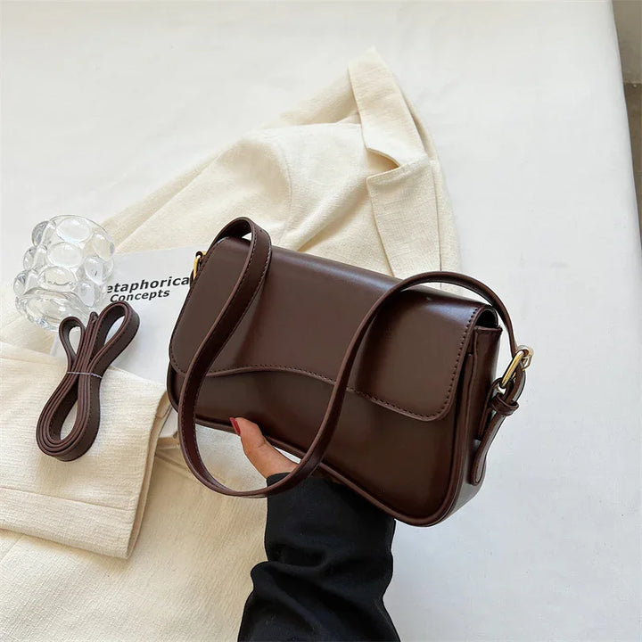 Brown leather crossbody bag with adjustable strap held over cream blazer and book on white surface