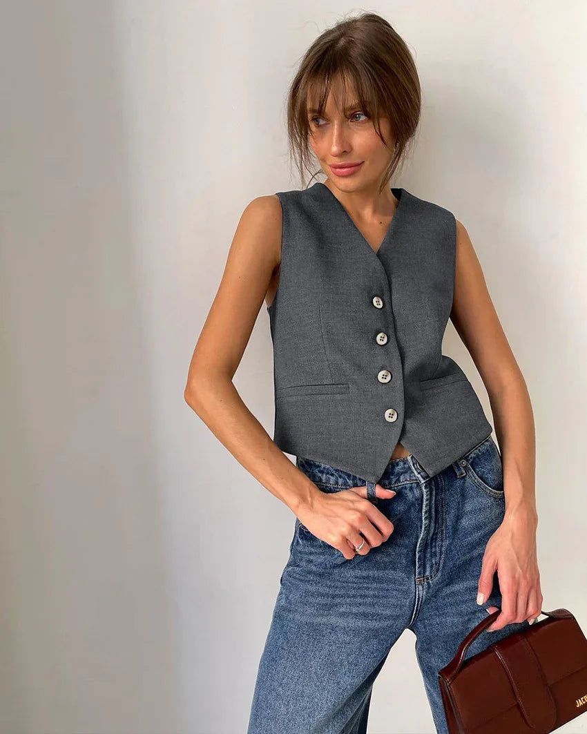 Woman wearing sleeveless gray buttoned vest with blue jeans holding brown leather handbag against plain background
