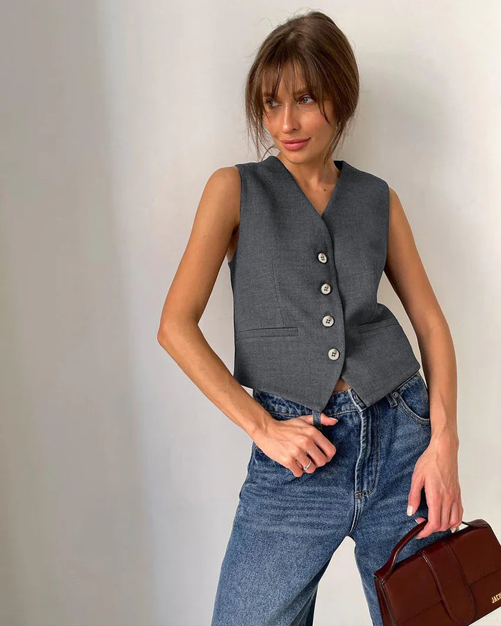 Woman wearing sleeveless gray buttoned vest with blue jeans holding brown leather handbag against plain background