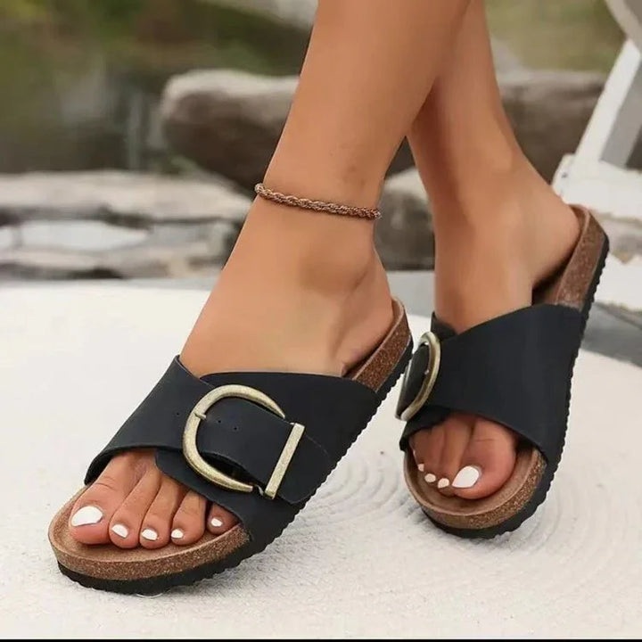 Close-up of feet wearing black slide sandals with large gold buckle and cork footbed outdoors