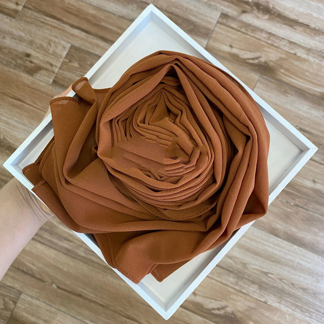Hand holding white tray with neatly rolled rust-colored chiffon fabric on wood floor background