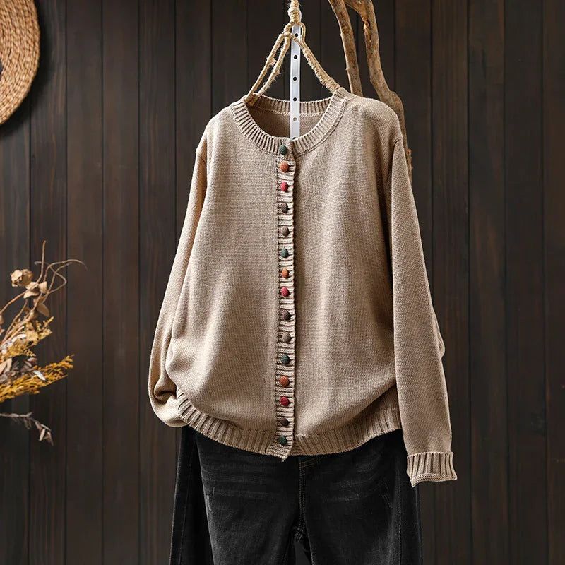 Beige knitted cardigan with colorful round buttons hanging on wooden hanger against dark wooden wall