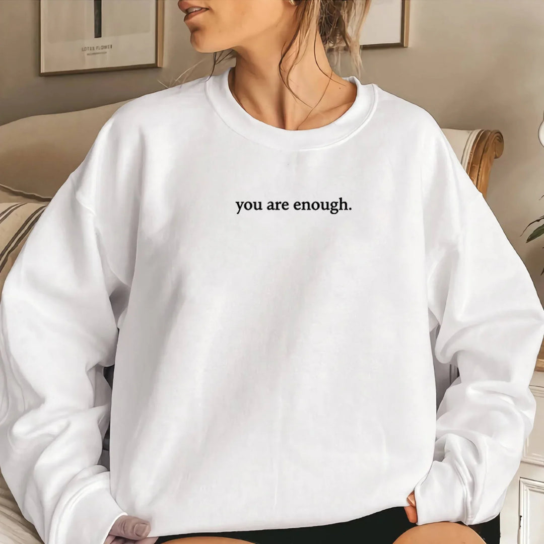 Woman wearing white sweatshirt with inspirational text 'you are enough' in cozy indoor setting