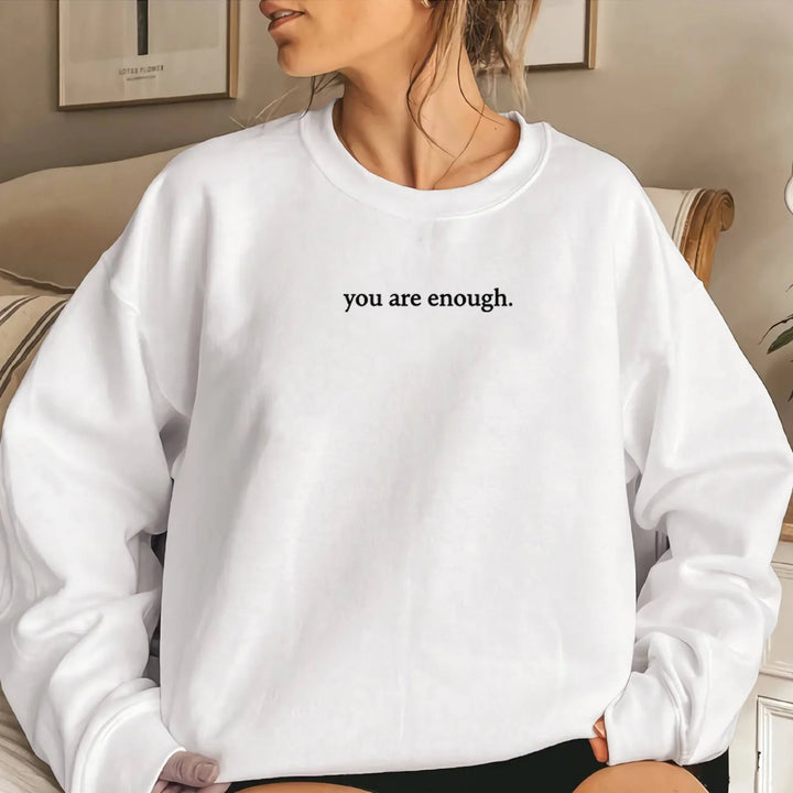 Woman wearing white sweatshirt with inspirational text 'you are enough' in cozy indoor setting