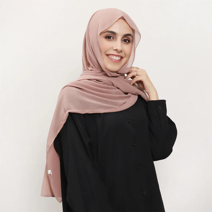 Smiling woman wearing a dusty pink hijab and black button-up top against white background