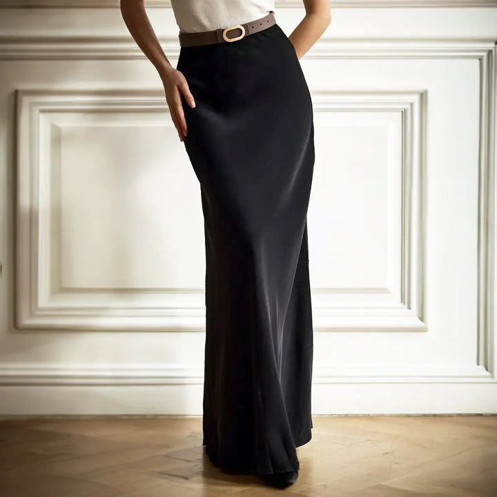 Woman wearing a high-waisted black satin maxi skirt with a brown belt, standing indoors on wooden floor