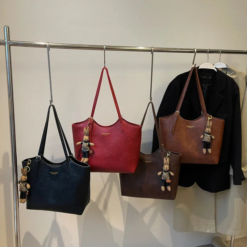 Black, red, and brown leather tote bags with bunny keychains hanging on a metal rack with coats in the background
