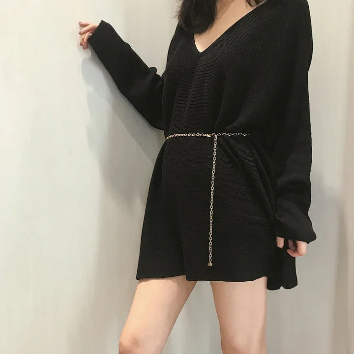 Woman wearing black oversized sweater dress with gold chain belt against plain wall