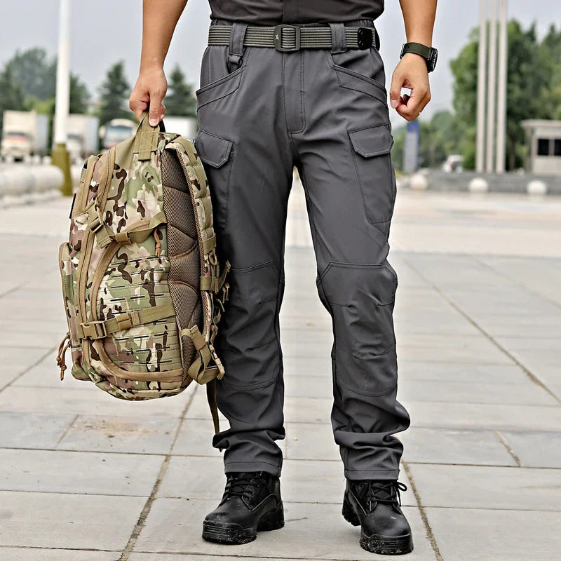 Man wearing gray tactical pants and black boots holding a camouflage military backpack outdoors