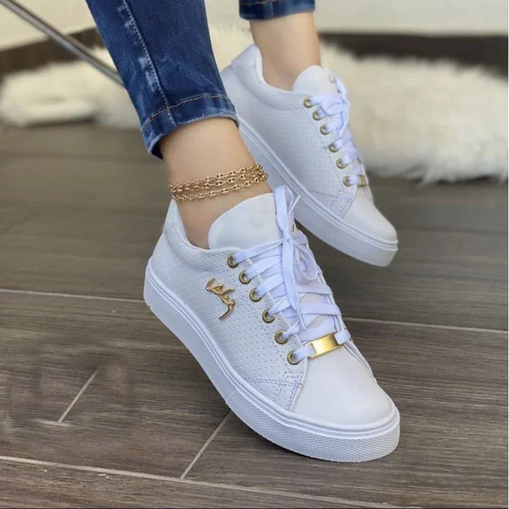 White perforated sneakers with gold eyelets and logo, worn with rolled blue jeans and gold anklet on wooden floor