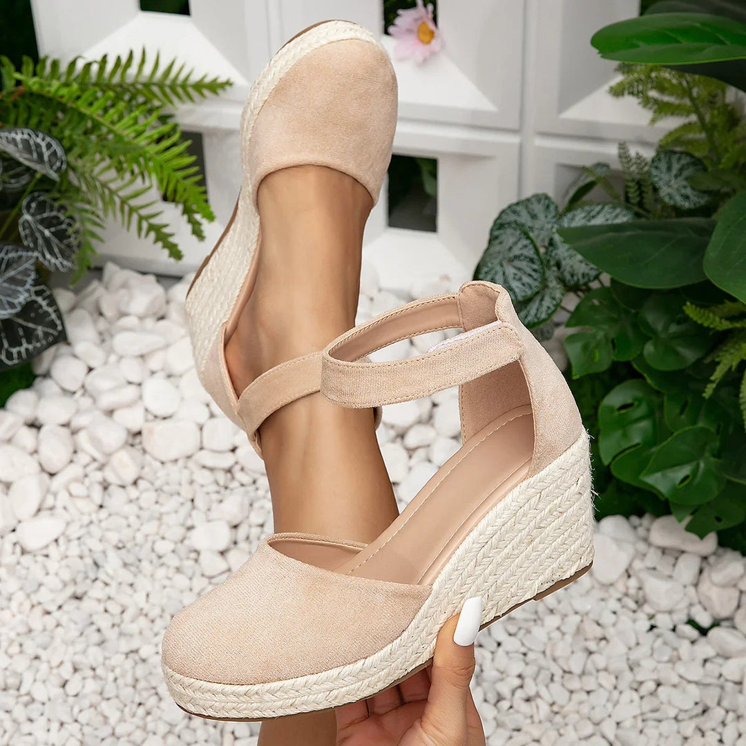 Beige espadrille wedge sandals with ankle strap on white pebbles and green plants