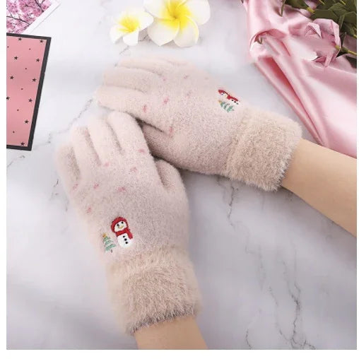 Soft beige winter gloves with snowman embroidery worn on hands, placed on marble surface with flowers and pink fabric