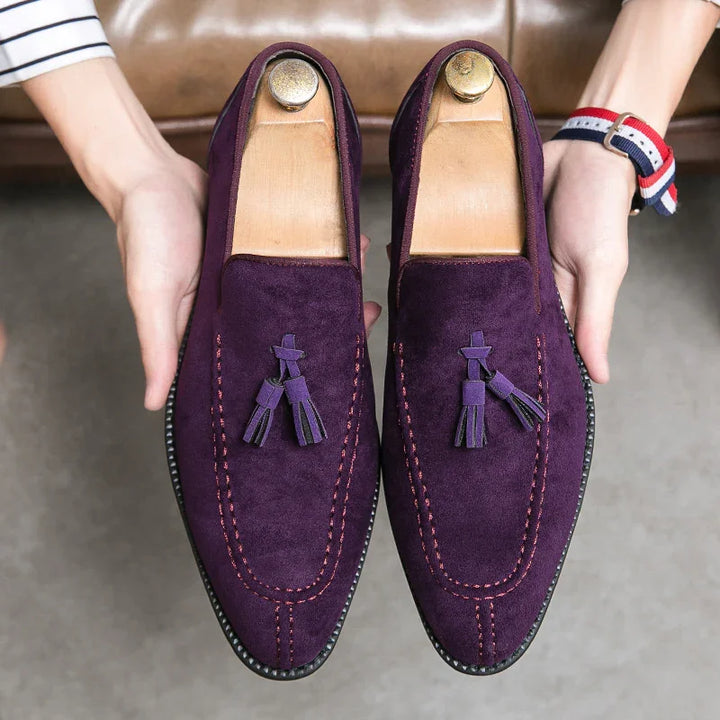 Pair of purple suede men's tassel loafers with pink stitching held above gray floor